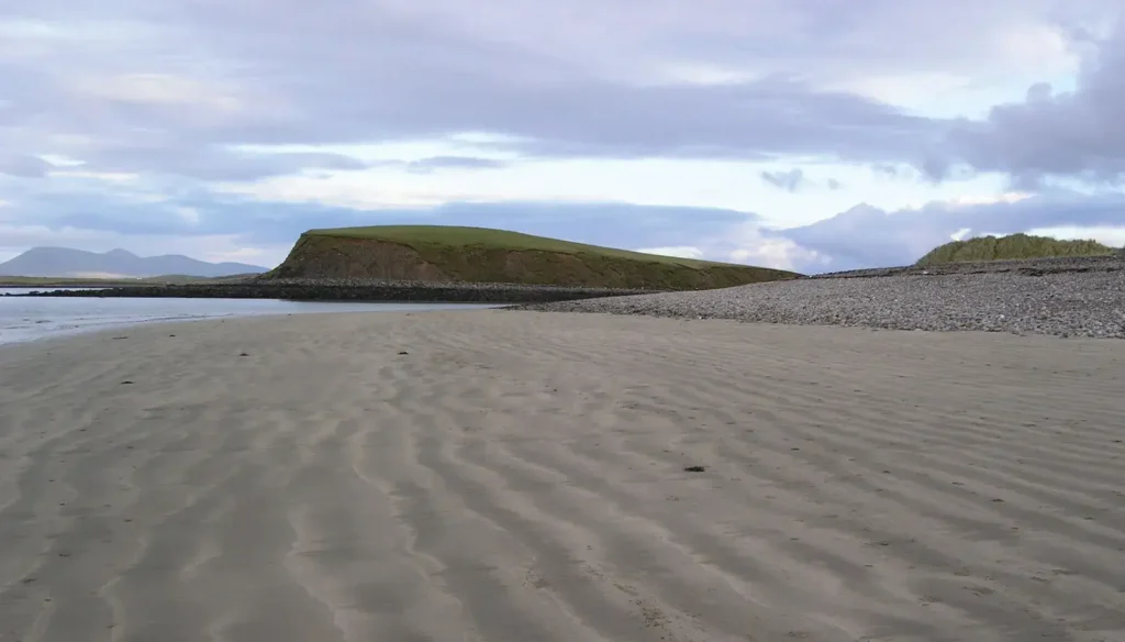 Playa de arena con una colina cubierta de hierba bajo un cielo nublado (drumlin)