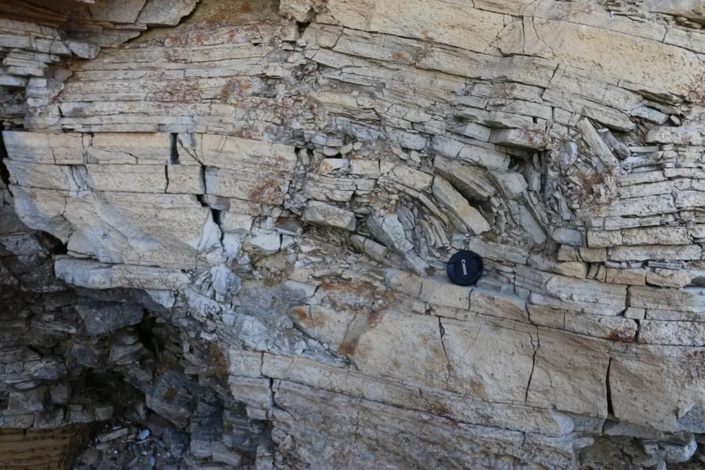 Capas de roca sedimentaria plegadas junto a una tapa de objetivo de cámara (pliegue de flexión de falla)