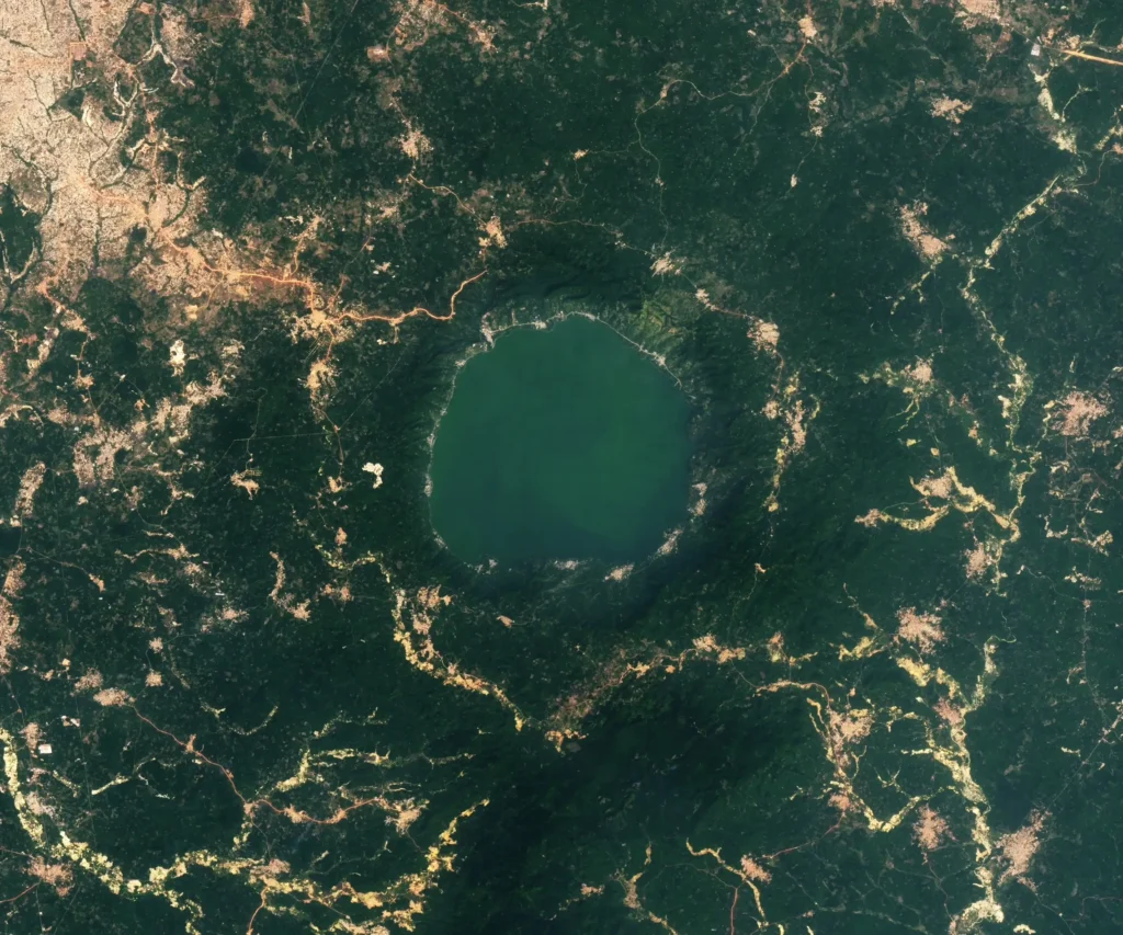 Landsat satellite image of Lake Bosumtwi in Ghana, a circular lake roughly 8 km across set in dark forested terrain with gold-mining scars along the rim