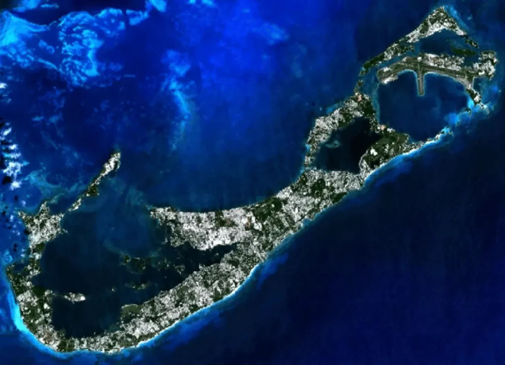Satellite view of Bermuda from orbit, a small hook of land surrounded by a pale ring of shallow reef in the deep blue open Atlantic.