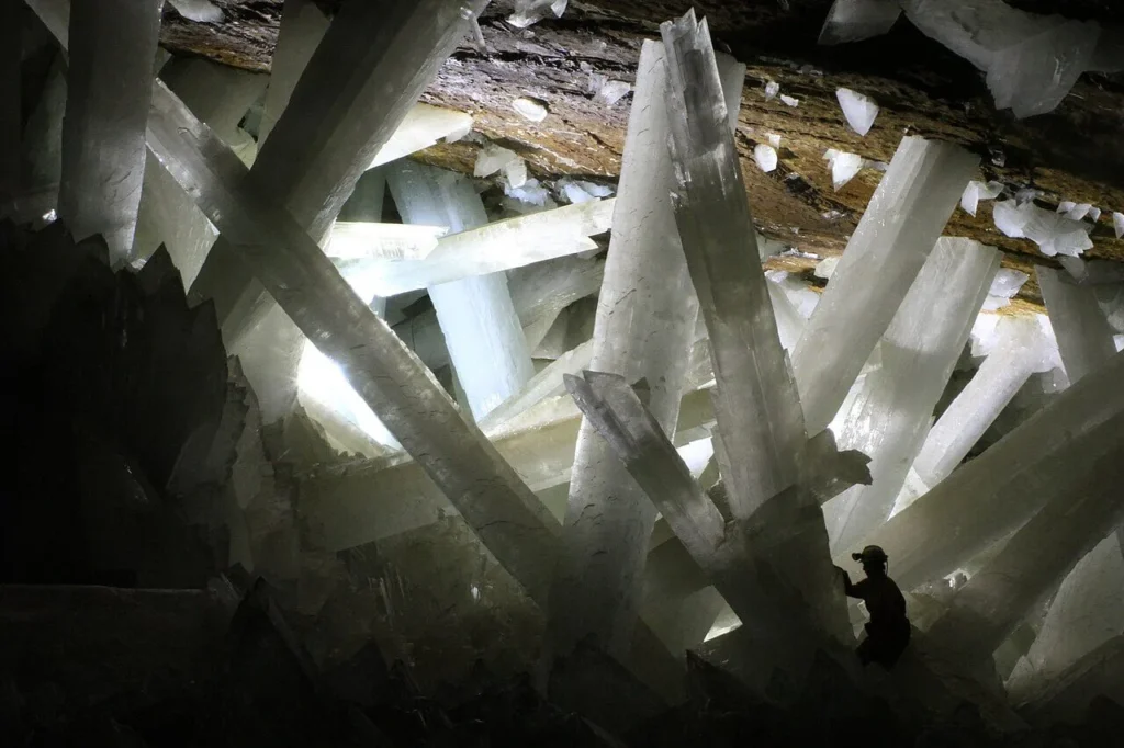 A person in protective gear dwarfed by massive translucent selenite gypsum beams crossing the Cave of Crystals at Naica, Mexico