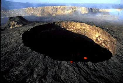 Helicopter view of the active lava lake inside Erta Ale's summit crater in February 1994, with two red-suited volcanologists standing on the crater rim for scale and red molten lava breaking through the dark crust below