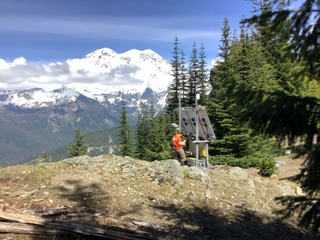 A USGS geophysicist crouched beside an equipment box at remote monitoring station PR05 on the flank of Mount Rainier, with the snow-covered summit of the volcano visible in the background.