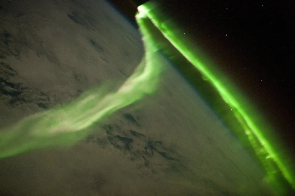 Sinuous green aurora australis photographed from the International Space Station above the southern Indian Ocean, with cloud deck below and Earth's atmospheric limb visible on the horizon
