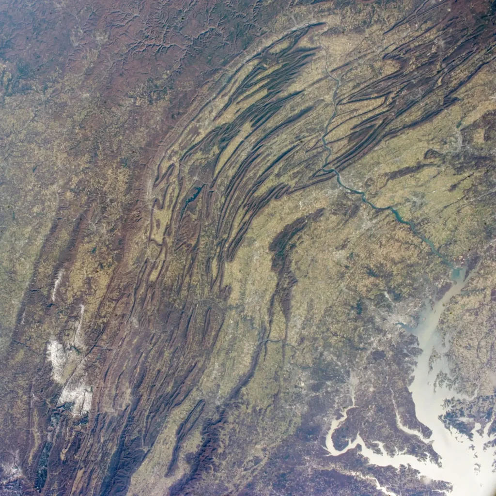 Oblique view from low Earth orbit showing the parallel forested ridges and farmed valleys of the Appalachian Mountains stretching from Pennsylvania southwest into West Virginia, with the Chesapeake Bay reflecting sunlight in the distance