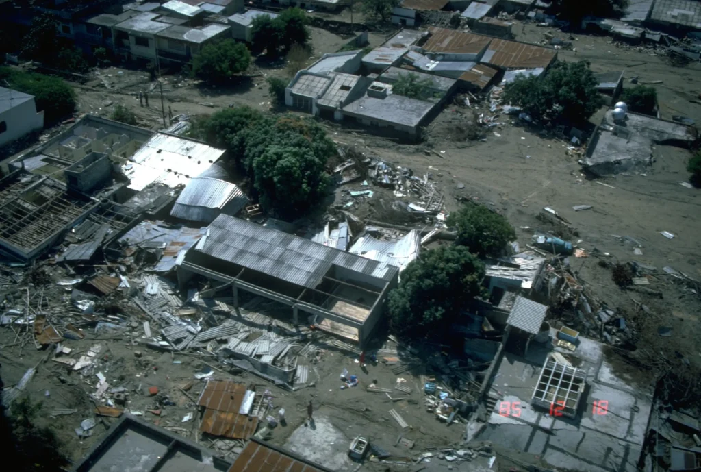 A wide aerial view of the town of Armero, Colombia, after the 13 November 1985 Nevado del Ruiz lahar, showing a flat expanse of grey mud, debris and ruined structures where the town once stood.