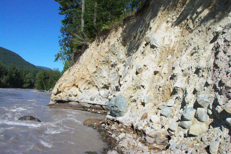An eight-metre-thick vertical outcrop of the Osceola Mudflow exposed in a river cut fifty kilometres downstream from Mount Rainier, showing a chaotic clay-rich matrix with angular cobbles and visible normal grading toward the base.