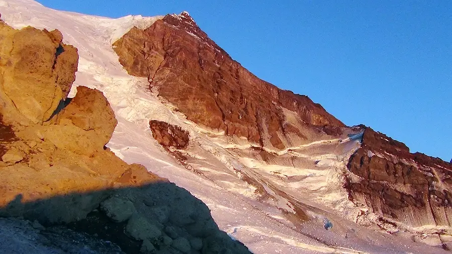 The west flank of Mount Rainier photographed at sunset from St. Andrew's Rock monitoring station, showing the Sunset Amphitheater, Tahoma Glacier headwall and steep upper slopes of the volcano lit in warm light against a deep blue sky.