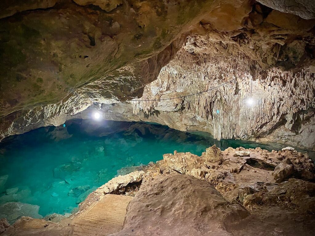 A blue-water cenote in Homun, Yucatán, a flooded limestone sinkhole with clear turquoise water beneath a rock overhang.