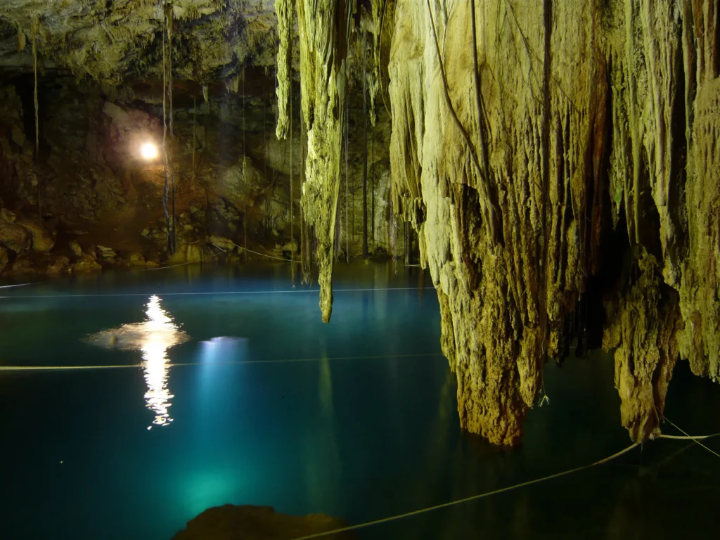 Interior of a Yucatán cenote, a limestone cavern with a pool of clear blue freshwater lit by a shaft of daylight from above