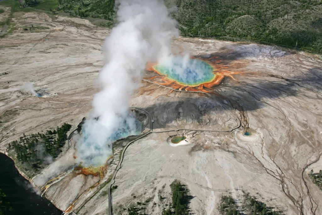 Aerial view of the bright blue Grand Prismatic Spring and the steaming crater of Excelsior Geyser in Yellowstone's Midway Geyser Basin, ringed by orange and yellow microbial mats.