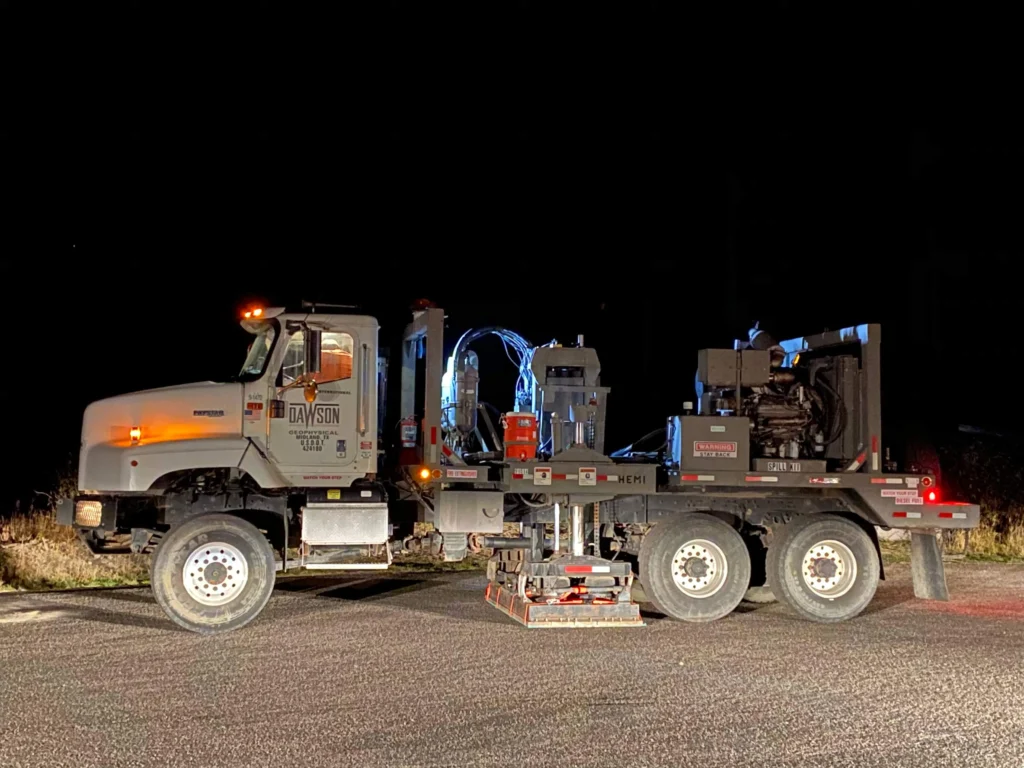 A 53,000-pound vibroseis truck parked at a roadside turnout in front of a Continental Divide sign in Yellowstone National Park at night, the sign lit by headlights.
