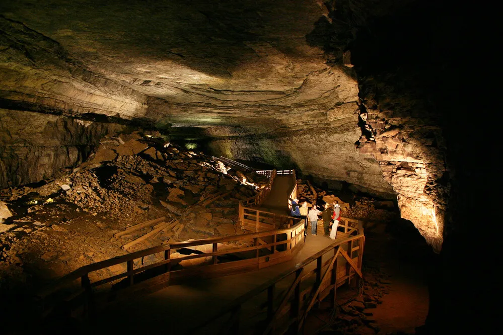 Visitors and a ranger on a boardwalk dwarfed by the vast dark tunnel of Broadway, the Main Cave passage inside Mammoth Cave.