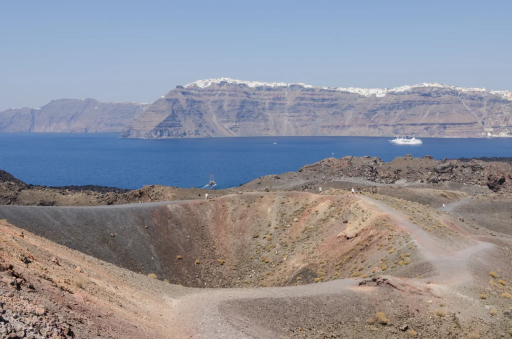 The dark volcanic island of Nea Kameni rising from the blue water of the Santorini caldera, with the caldera cliffs in the background