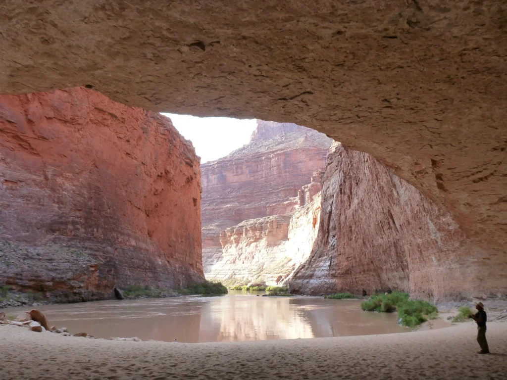 A vast arched cavern mouth opening onto bright daylight and a river beyond, the cave floor and ceiling dwarfing the view out.