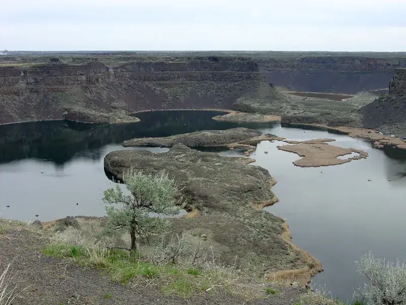 Ground-level photo of Dry Falls in Washington: a curving cliff of dark basalt roughly 400 feet high and 3.5 miles wide, with small lakes pooled at its base.