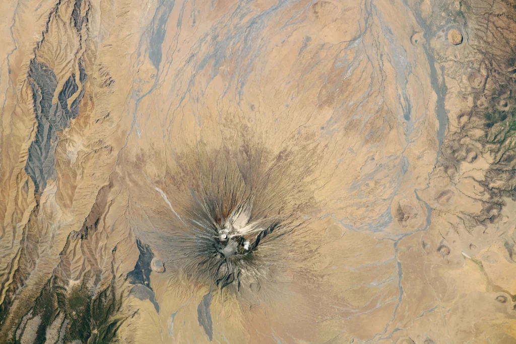 Astronaut photograph of the symmetric cone of Ol Doinyo Lengai volcano in Tanzania, with a pale ash-capped summit and light-colored lava flows streaking its flanks.