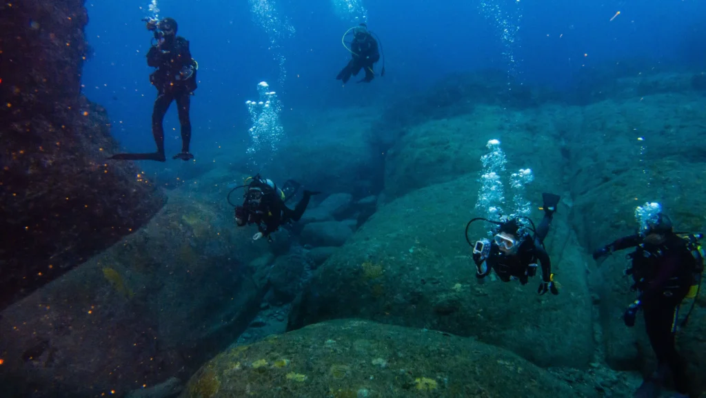 Five scuba divers above the Yonaguni Monument, where a straight channel runs between blocky sandstone terraces