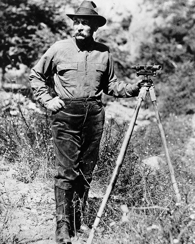 William Mulholland standing beside a surveyor’s transit on a tripod in the field